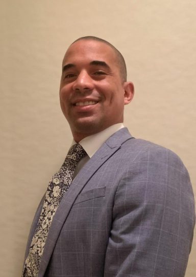 Benjamin Hare Man smiling in a tailored suit, posed slightly to the side against a light background.